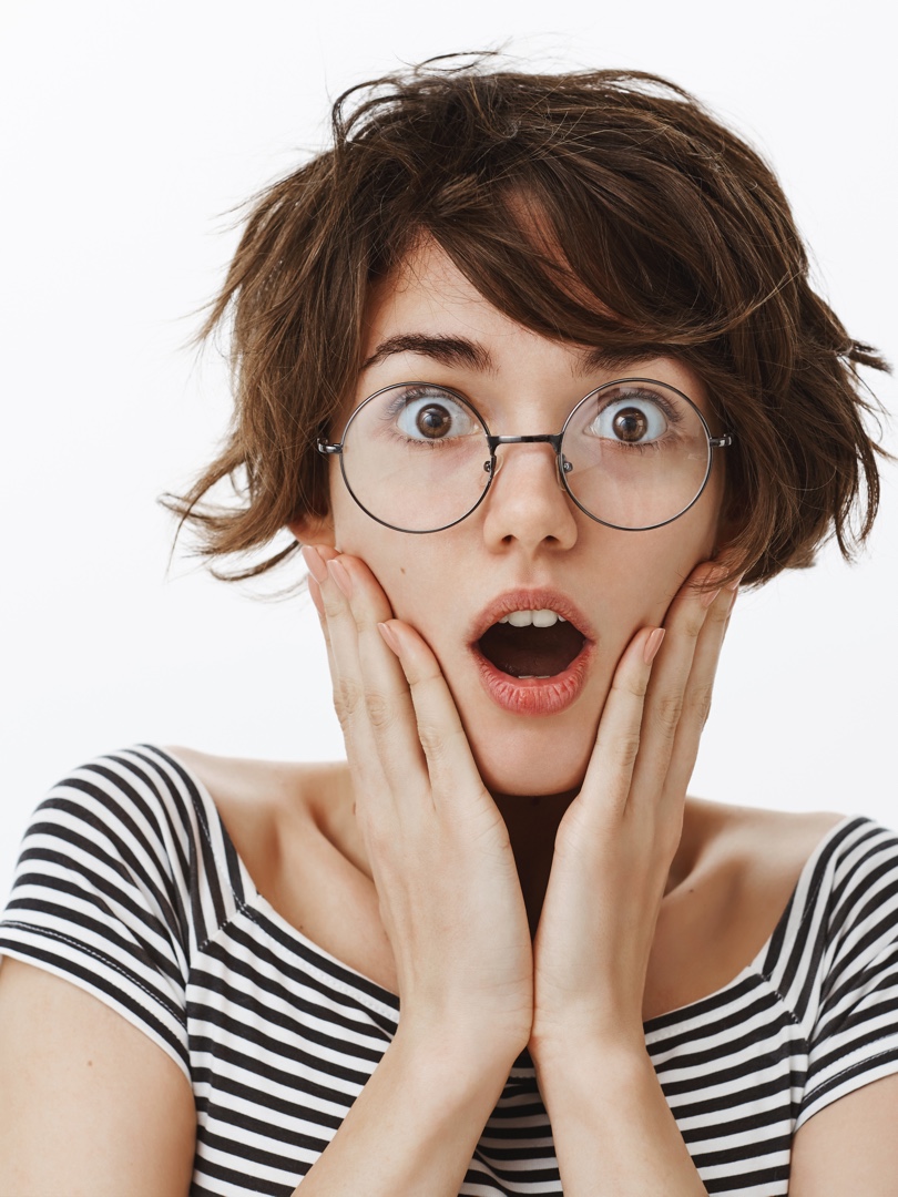 Impressed and shocked charming female with stylish short haircut in round trendy glasses, gasping and dropping jaw, holding palms on cheeks, being stunned with news or rumors spread across office